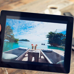 Man holding digital tablet with vacation photograph. He could be searching a travel website for hotels or resorts. Home interior setting with a wood textured table in the background. There is also coffee and a notebook in the background.